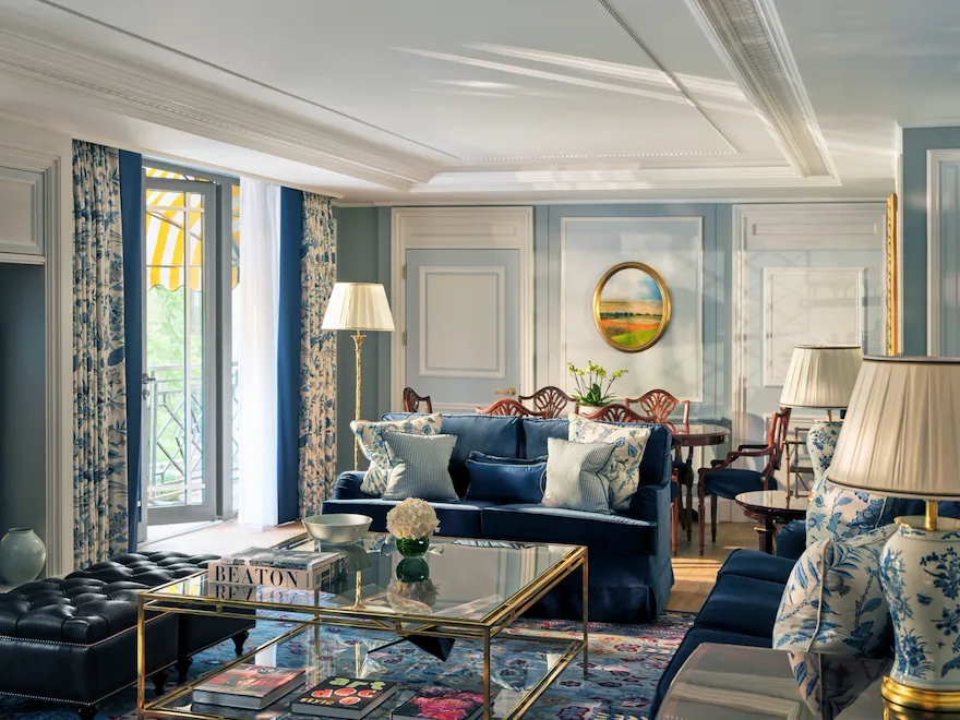 The Hyde Park Suite living area at The Dorchester, showcasing soft blue interiors, plush seating, decorative panelling, a glass coffee table and French doors opening onto a balcony with views of Hyde Park.
