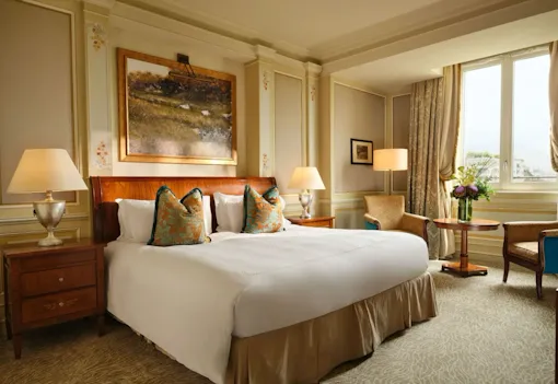 The room is decorated in soft gold colours and features decorative gold and blue cushions. There is a large painting above the bed and a large window that lets in plenty of natural light.