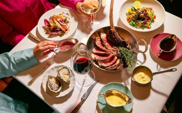 Dining table at The Grill at The Dorchester, showcasing a sliced Tomahawk steak served on the bone, accompanied by oysters, seasonal salads, rich sauces and glasses of red wine in an elegant sharing-style feast.