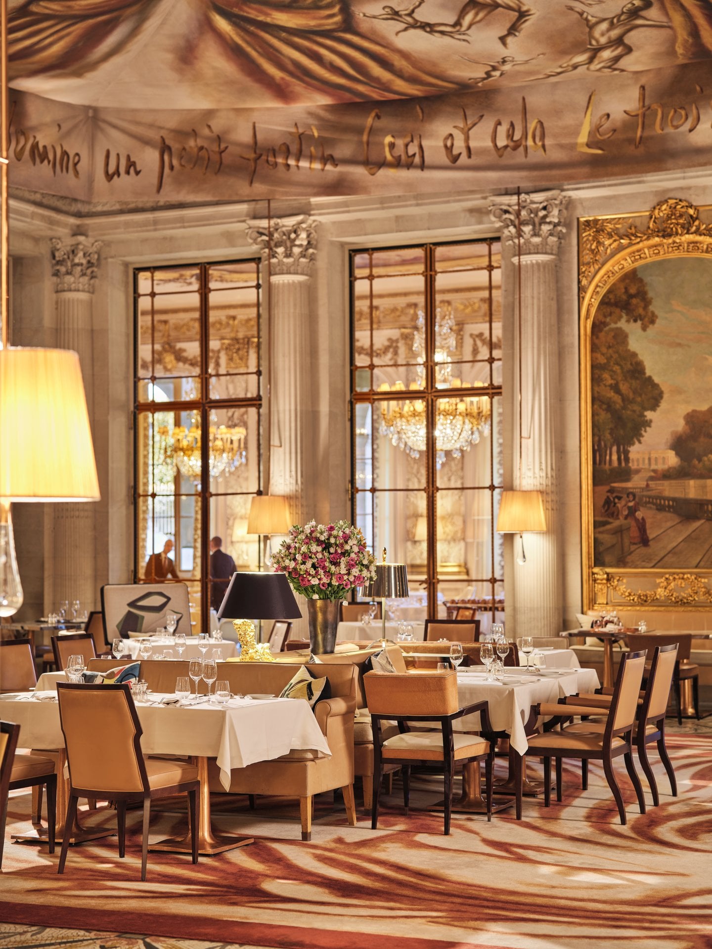 Elegant dining room with large windows, chandeliers, floral centerpiece, and polished decor.