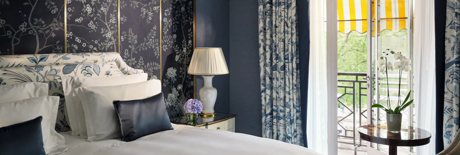 Bedroom in The Hyde Park Suite at The Dorchester, featuring blue floral décor, a king-sized bed and a private balcony with a yellow-striped awning.