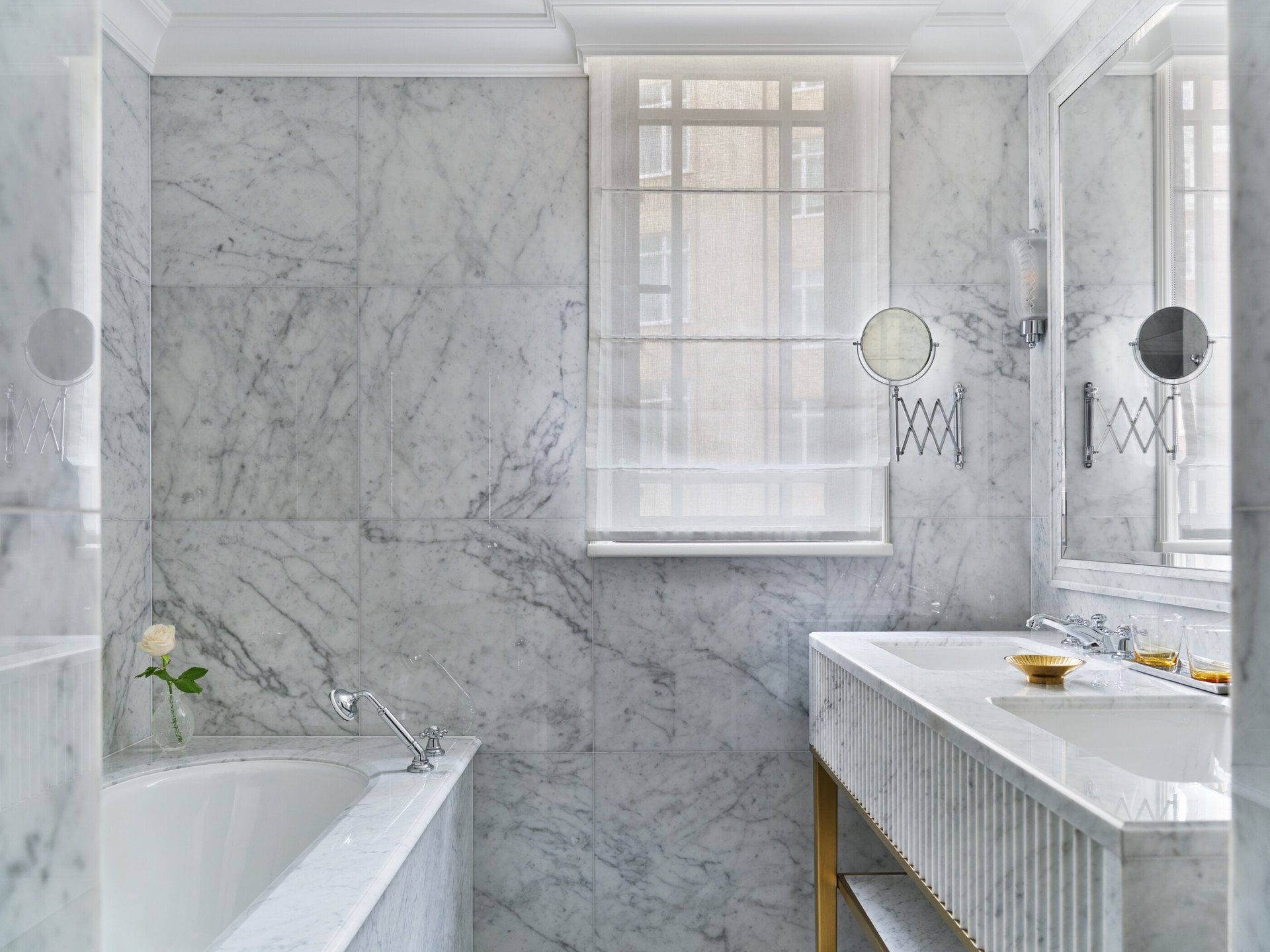 The Superior Room bathroom at The Dorchester, showcasing floor-to-ceiling white marble, a built-in bathtub, a fluted marble double vanity and a large mirror that enhances the bright, airy space.