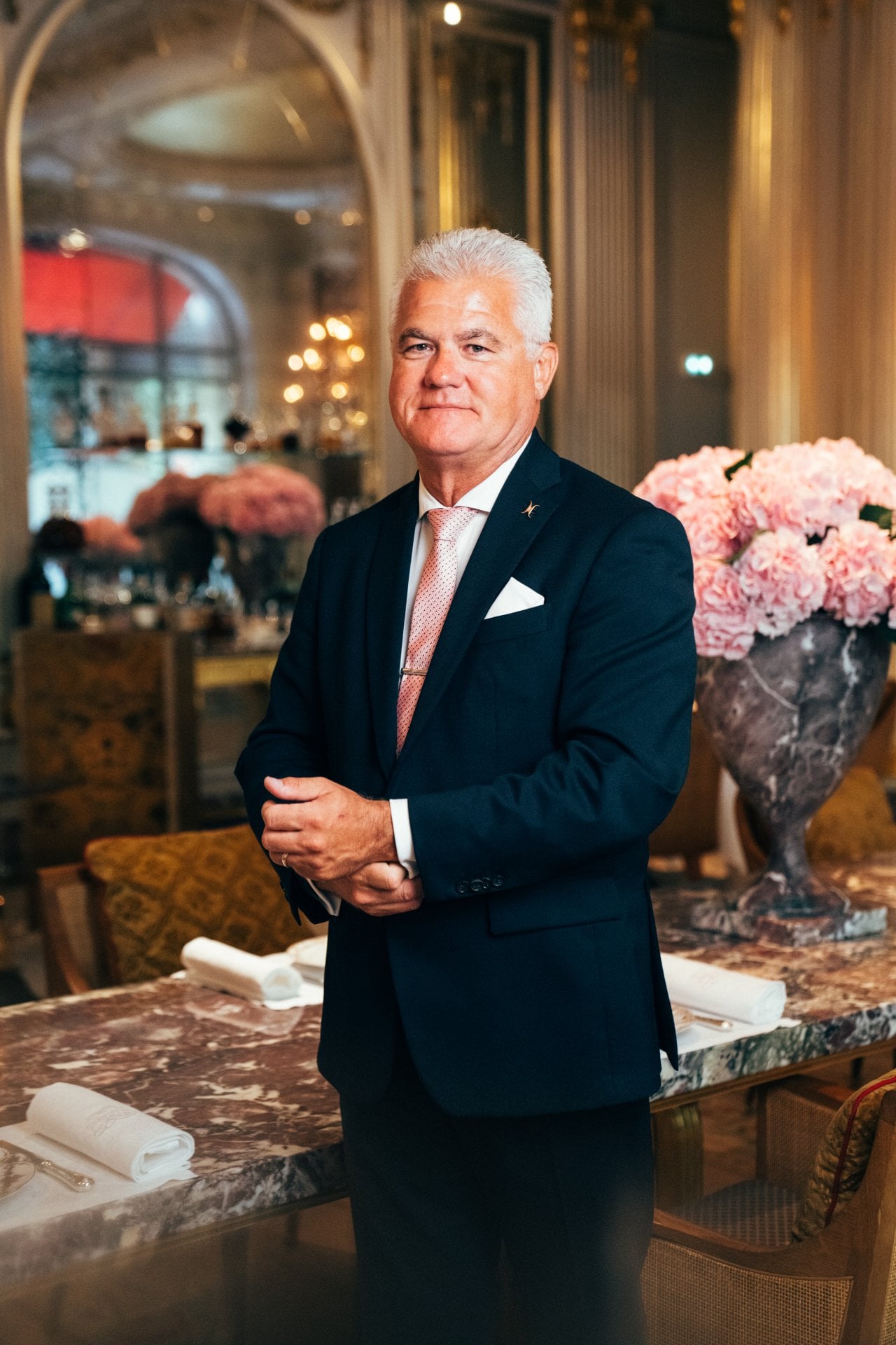 Person in formal suit standing in elegant room with pink flower arrangement and marble table.