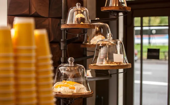 Display of freshly prepared pastries at Parcafé at The Dorchester, showcasing sweet treats presented under glass cloches on tiered shelves, with takeaway coffee cups and views towards Hyde Park visible through the café window.
