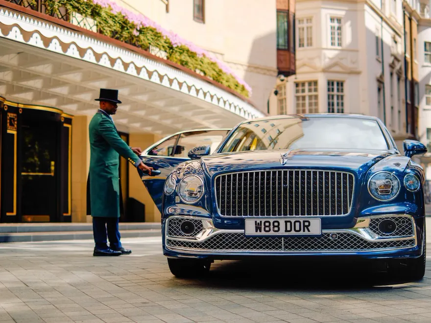 The Dorchester Bentley positioned outside the hotel's main entrance on Park Lane, showcasing its distinctive chrome grille and personalised registration plate, with The Dorchester's elegant façade and entrance canopy in the background.