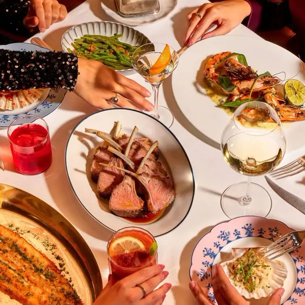 Dining table at The Grill at The Dorchester, showcasing a selection of signature dishes including sliced roast lamb, grilled prawns, dover sole, green beans, artisan bread and cocktails, presented for sharing.