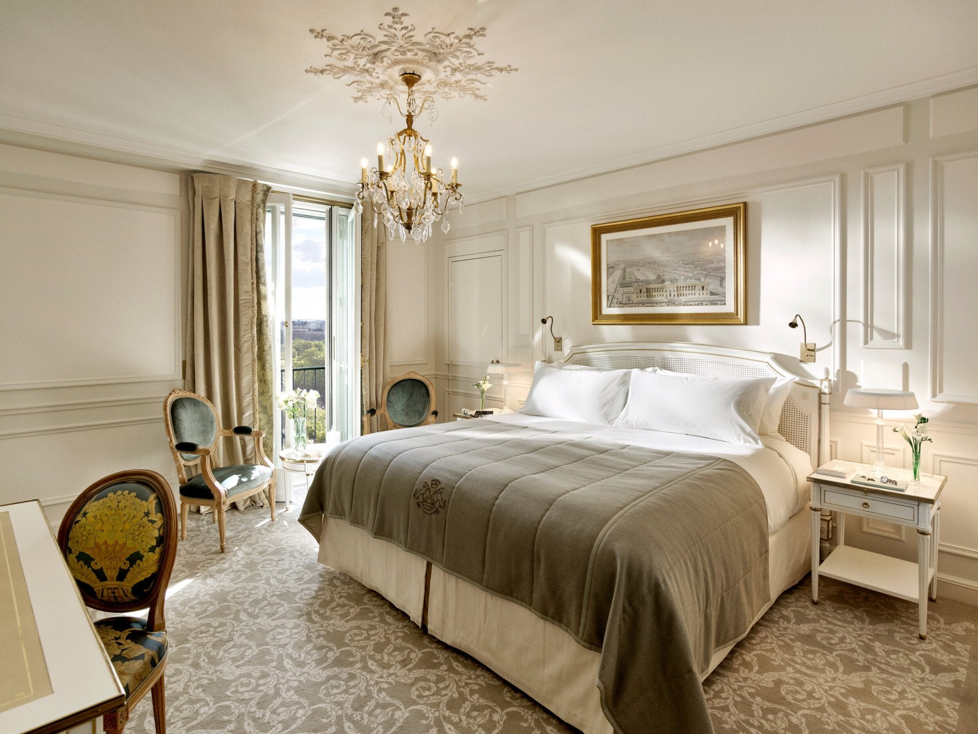 Global view of an executive room highlighting the light grey and cream undertones, as well as the view over the Tuileries gardens in the background. Jewelry and books are on the bedside tablesAt Le Meurice, Paris.