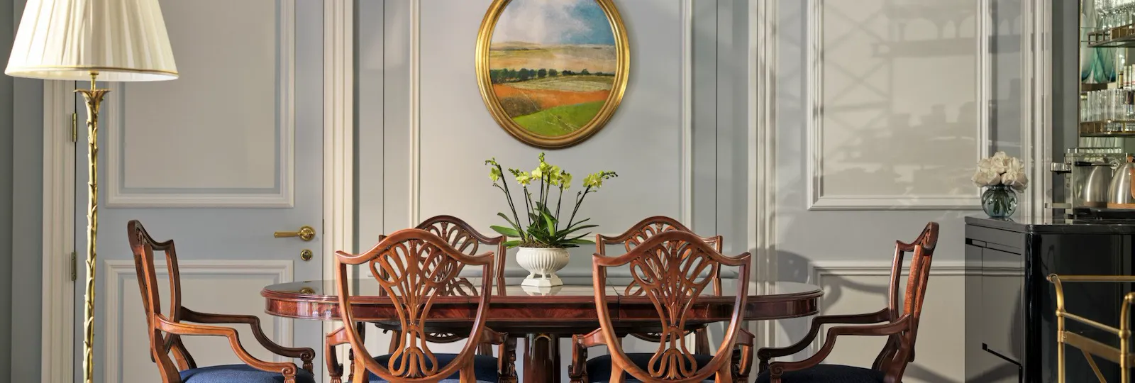 The Hyde Park Suite at The Dorchester, showcasing a formal dining area with a polished wooden table, blue-upholstered dining chairs, decorative wall panelling and an oval-framed landscape painting.