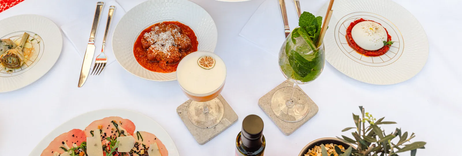 Two woman sitting on a red bench sharing a pizza, cocktails and Italian dishes are displayed on a white table.