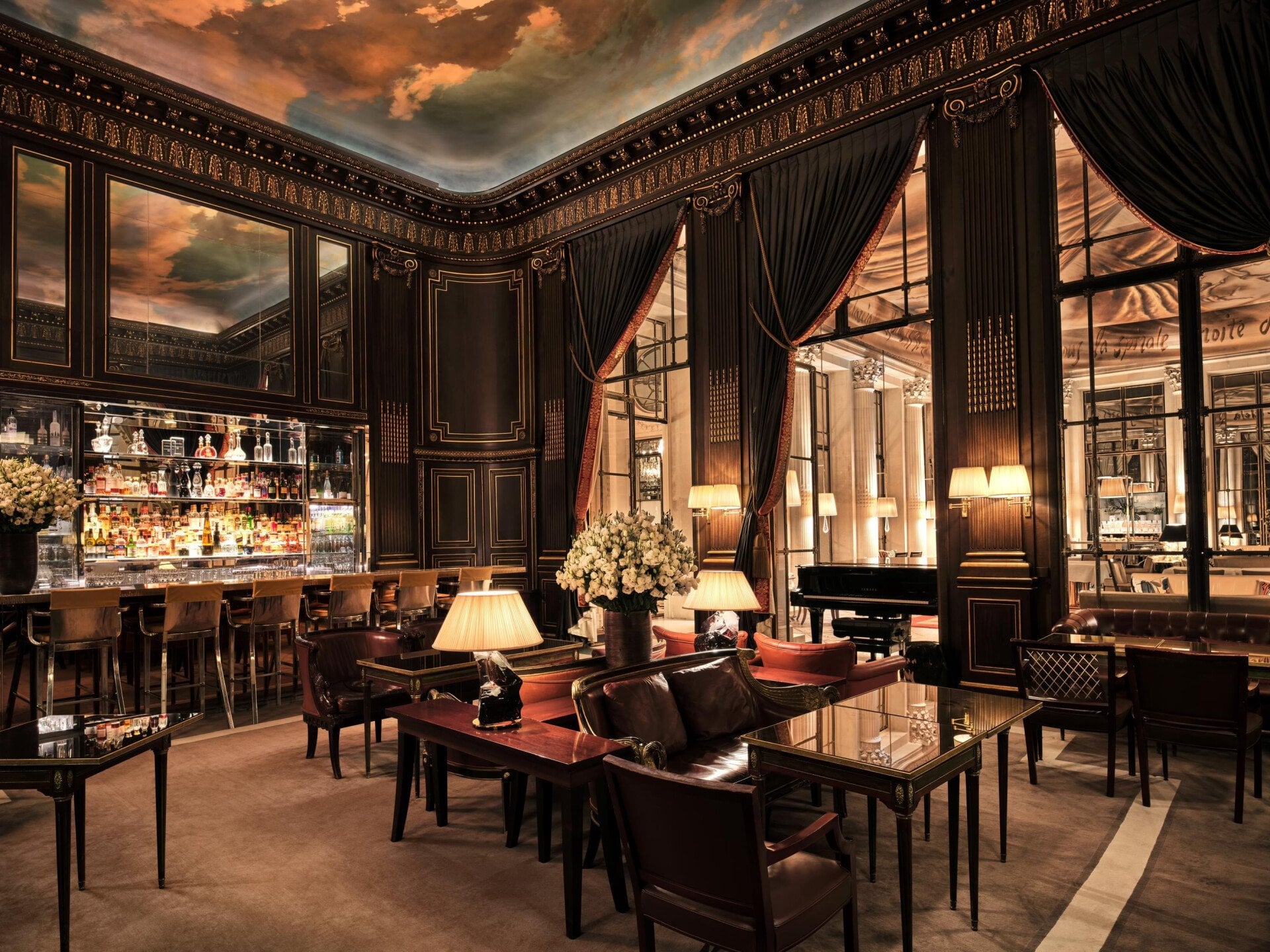 In the center of the photo, we see lamps lit on tables with armchairs and dark red cushions, and a bouquet of white flowers in a vase. On the left is bar 228 with high chairs. We can see the bottles on the shelves. Above the bar is a mirror reflecting the ceiling, which represents a sky with clouds. We look at the woodwork on the walls, with its gilded details. To the right is a piano and large windows with red curtains, revealing another room at Le Meurice, Paris.
